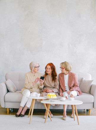 Three senior women enjoying tea and conversation indoors, looking at a smartphone together.